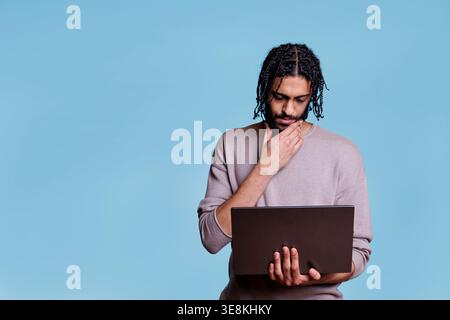 Young thoughtful arab man holding laptop while analyzing software code and rubbing chin. Pensive person thinking in doubt while reading business report on portable computer Stock Photo