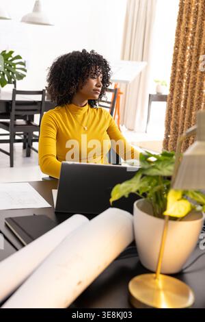 Adult woman in mustard top sitting at home desk reviewing rolled plans and typing on laptop Stock Photo