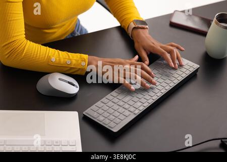 Female mustard yellow top typing on slim gray wireless keyboard at matte black desk with smartwatch Stock Photo