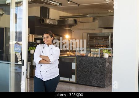 Woman chef in white jacket leaning, smiling at bakery door by glass OPEN sign, copy space Stock Photo
