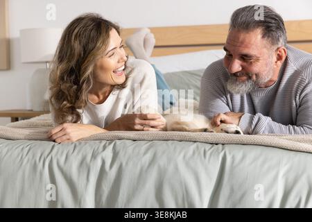Couple mid-adult woman and mature adult man lying on bed at home, laughing and petting dog Stock Photo