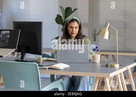 Woman in green sweater, headband working on laptop at coworking table, viewing floor plan monitor Stock Photo
