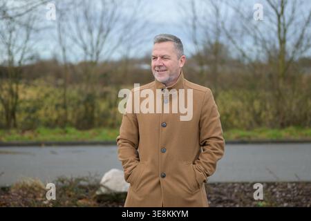 A smiling middle aged man wearing a brown coat stands outdoors with his hands in his pockets while looking to the side. The natural background and sof Stock Photo
