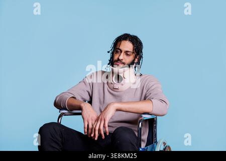 Exhausted arab man with neck brace sitting in wheelchair, healing from accident. Tired young patient wearing cervical collar protector for spinal injury recovering from trauma Stock Photo