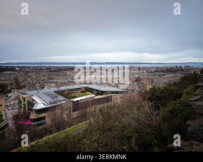 Wide panoramic view over Edinburgh with rooftops, modern architecture, harbor and moody winter sky from a hilltop. Stock Photo