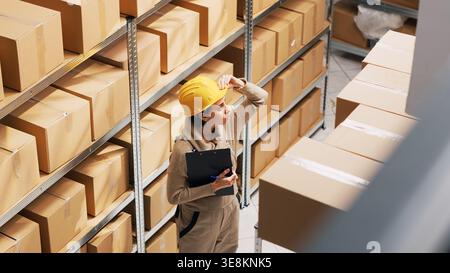 Female employee counting number of packages in depot storage room, checking products on shelves. Young woman working on stock logistics, doing inventory before products shipment. Stock Photo