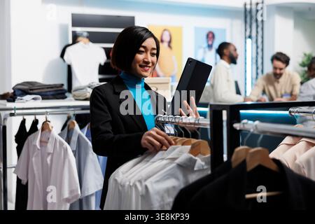 Clothing store smiling manager using digital tablet to check stock availability while browsing through apparel rack. Shopping center boutique asian woman employee managing inventory Stock Photo