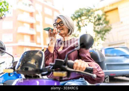 Mature woman riding scooter using smartphone voice assistant Stock Photo