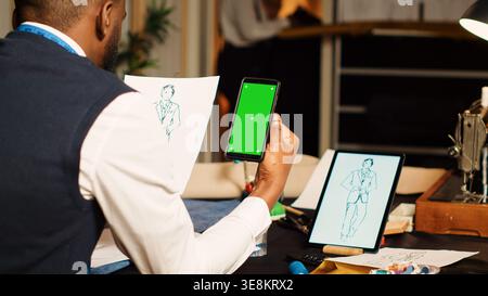 Tailor using greenscreen in atelier with team of designers, looking at isolated display on smartphone. Male couturier working with blank chroma key template on screen, tailoring concept. Stock Photo