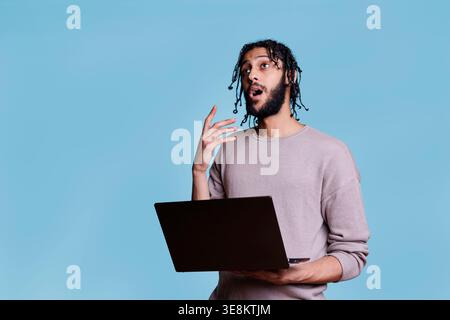 Puzzled arab man thinking about software project while holding laptop and looking upwards in doubt. Pensive person with thoughtful facial expression while coding on portable computer Stock Photo
