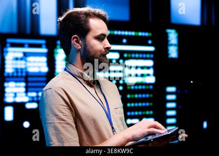 Focused serviceman checking functional blade servers in computer network security data hub. High tech facility infrastructure providing processing and memory resources for computational operations Stock Photo