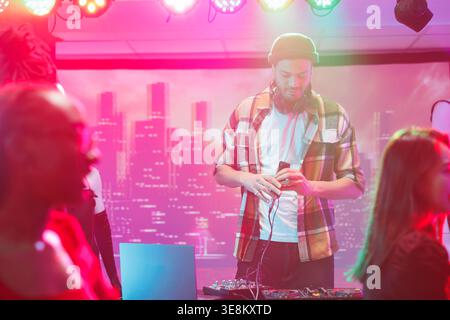 Young man musician using dj controller on stage during live concert in nightclub. Performer mixing electronic music tracks for clubbers at discotheque party in club at night Stock Photo