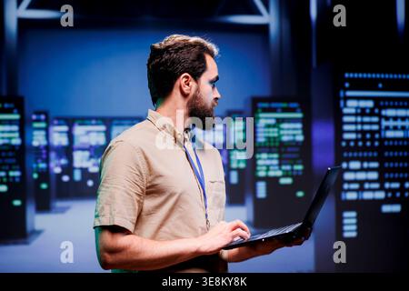 IT developer in server hub storing data management rigs, storage devices and networking devices offering vast amounts of computing resources. Employee doing infrastructure maintenance using laptop Stock Photo