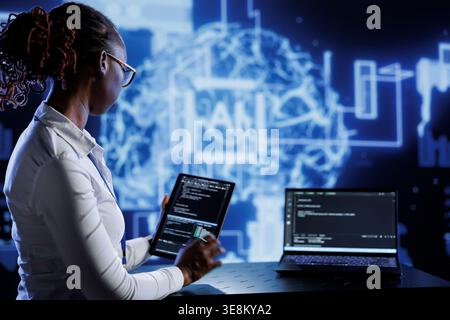 Computer scientist updates neural networks made of interconnected nodes, writing intricate binary code scripts on tablet and laptop. Server room expert uses programming to upgrade AI simulation model Stock Photo