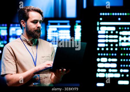 System administrator using laptop to check high tech server rigs. Rackmounts in computer network security data center establishment providing processing and memory resources needed for workloads Stock Photo