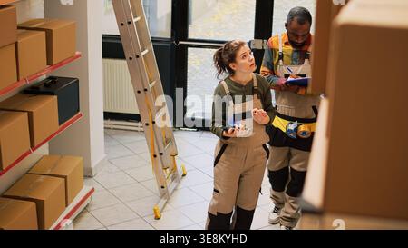 Warehouse diverse team reviewing orders and preparing products in packages for shipment, checking logistics on paper and tablet. Man and woman planning cargo distribution in storage room. Stock Photo