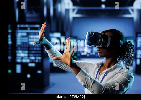 Tech support in data center uses virtual reality to prevent system overload. BIPOC VR technology user in server room ensuring enough network bandwidth for smooth operations Stock Photo