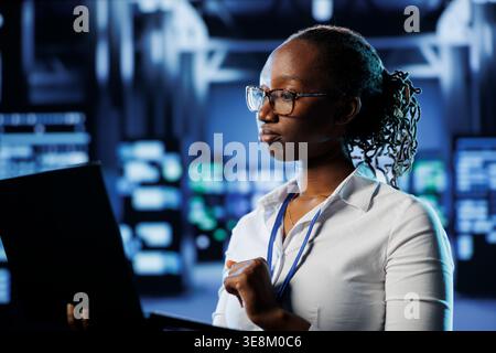 Proficient admin in high tech establishment uses laptop to prevent system overload. Competent expert between server cabinets ensuring enough network bandwidth for smooth operations Stock Photo