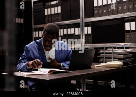African american manager reading management research, working overtime at bureaucracy record in storage room. Businessman analyzing administrative files, searching for accountancy report Stock Photo