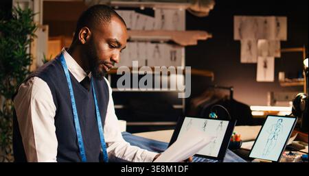 African american tailor checking sketches, using laptop and outlines on papers to compare fashion designs in atelier. Tailoring craftsman looking at clothing items models. Handheld shot. Stock Photo