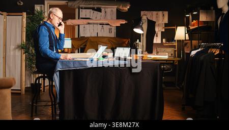 Craftsman calling client in workshop, ensuring perfect measurements and fabric on mobile phone call. Professional couturier designing handmade clothes and craft tailored items. Handheld shot. Stock Photo