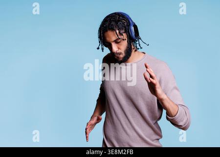 Concentrated arab man dancing while listening to music in headphones. Young person focused on songs playlist in wireless earphones moving hands and body on blue background Stock Photo