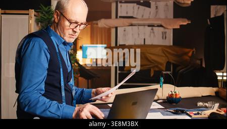 Craftsman looking for design on laptop, examining sketches with fashion items models. Experienced tailor designing clothes in luxury atelier, manufacturing handmade clothing line. Stock Photo