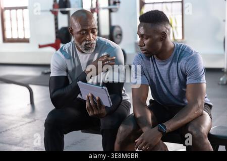 African American men, senior and adult, sitting in sportswear on padded bench, pointing at tablet Stock Photo