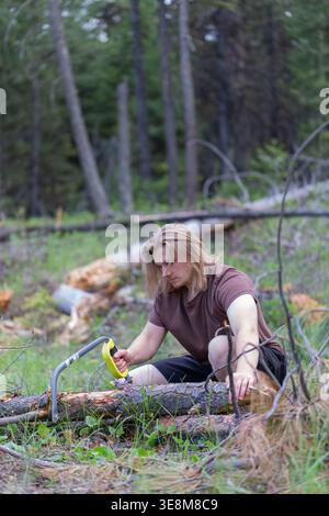 A young man with long blond hair crouches in a wooded area, using a bow saw to cut through a fallen log for firewood or shelter construction. Stock Photo