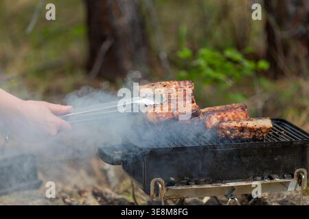 Close-up, dynamic shot showing several pieces of thick, pork ribs cooking on portable smoking grill. Hand uses metal tongs to manage the food. Stock Photo