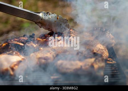 Close-up view of smoky chicken thighs sizzling on the grill while tongs handle the browned and charred pieces. Stock Photo
