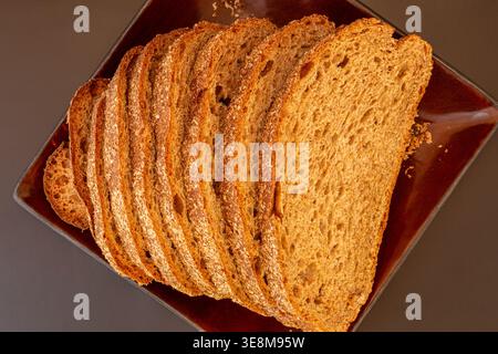 Slices of grain bread arranged on dark brown plate in top-down view revealing detailed texture of internal crumb and flaky bran crust. Dark background. Stock Photo