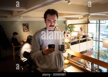 Handsome businessman enjoying coffee and scrolls smartphone after finishing work at co-working cafe. Stock Photo