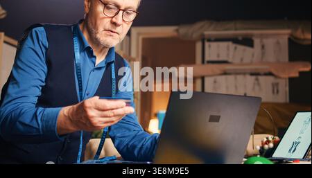 Old couturier shopping tools online, using credit card at laptop in workshop. Senior small business owner buying fabric or materials to create modern fashion items, tailoring. Handheld shot. Stock Photo