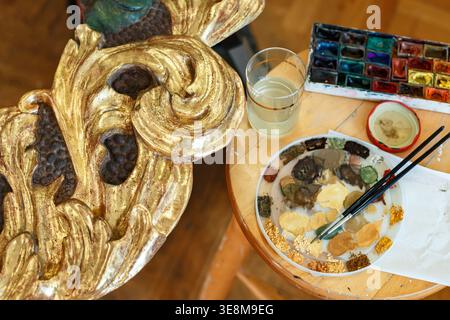 An artist sets up various paint colors, brushes, and a glass of water on a wooden table in a studio during the afternoon. Stock Photo