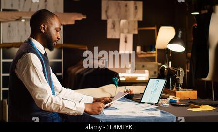 Fashion designer looks at sketches working with laptop to create modern clothing design in atelier. Tailor crafting new clothes collection in modern workshop, handmade concept. Stock Photo