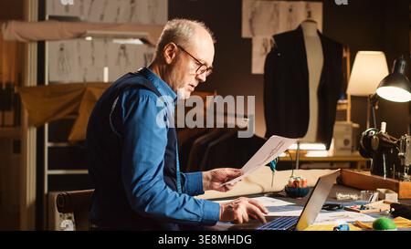 Senior couturier work on items design with sketches, using laptop to review material and custom made sizes. Male professional tailor creating luxury clothes in workshop atelier. Stock Photo