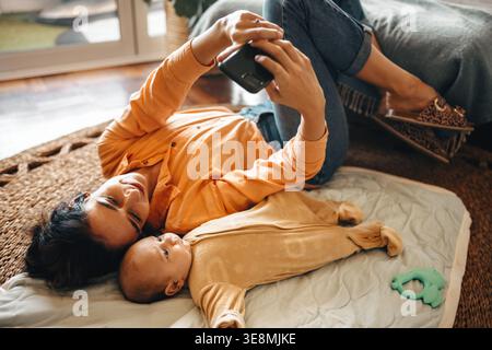A happy mother lies on a mat with her baby son, smiling and holding a phone to take a selfie. Captures love, bonding, and the joy of motherhood in a c Stock Photo