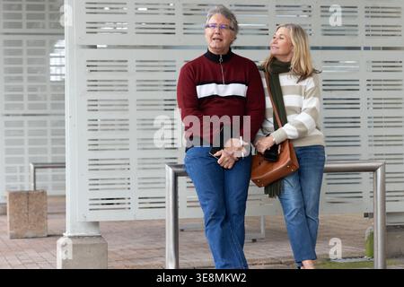 Senior couple standing and leaning against metal railing at plaza, wearing sweaters and holding bag. Elderly, duo, barrier, urban, sheltered, brickwork, scarf Stock Photo