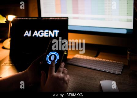 Individual interacts with smartphone featuring biometric security while seated at a desk with multiple computer screens showing data and information Stock Photo