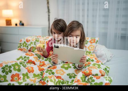 Two young sisters using a digital tablet in bed, one holding a green apple Stock Photo