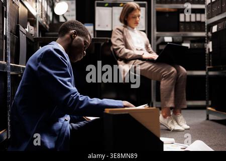 Overworked inspectors checking case files in arhive room, working late at night at criminal investigations. Tired private detectives analyzing missing person report, discussing crime scene evidence Stock Photo