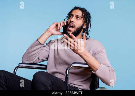 Emotional irritated arab man with disability talking on smartphone while sitting in wheelchair. Angry person with physical imparment making gestures while chatting on mobile phone Stock Photo