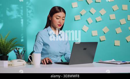 Sleepy asian office clerk woman slowly typing data after pulling all-nighter. Modern bright colourful office desk in professional administration workplace over blue studio background Stock Photo
