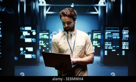 System administrator walking between operational server racks in computer network security data center, ensuring optimal performance. Engineer monitoring energy consumption across components Stock Photo