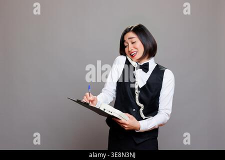 Smiling asian receptionist chatting with client on landline phone and writing in clipboard. Young attractive waitress in uniform having friendly conversation on retro telephone Stock Photo