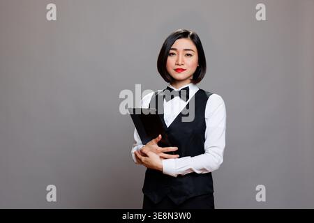 Confident young asian woman waitress wearing uniform and bow tie standing with digital tablet portrait. Serious receptionist holding portable gadget in hands and looking at camera Stock Photo