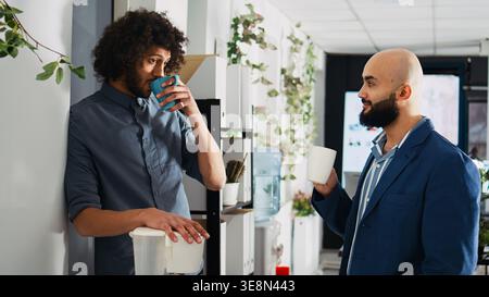 Employees relaxing on coffee break, laughing together and having fun before starting to work on small business. Two partners talking to each other, standing in coworking space. Stock Photo