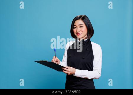 Smiling asian waitress wearing uniform holding clipboard, writing with pen and looking at camera. Cheerful woman receptionist taking notes studio portrait on blue background Stock Photo