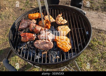 Mixed meat grilling on charcoal barbecue. Close up of assorted meat cooking on a charcoal grill outdoors, with tongs turning marinated pieces during a Stock Photo